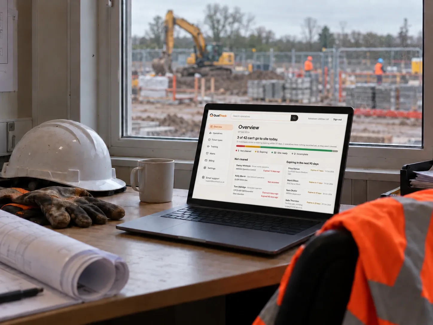 A laptop on a site office desk showing the QualTrack Overview, with a helmet, gloves and drawings beside it and the site through the window.